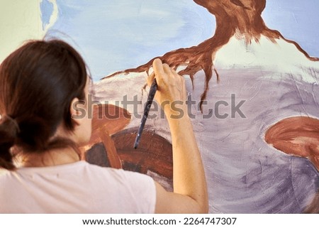 Girl artist hand holds paint brush and draws abstract nature landscape on white canvas at outdoor art painting festival, paintings art picture process. Woman artist paints atmospheric surreal picture