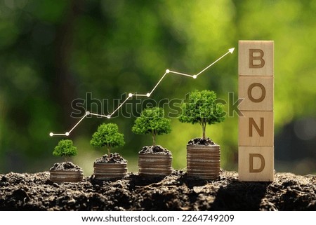The tree grows on a pile of coins and a wooden cube in the word Bond with green background. Investment on bonds concept. Raising funds to fund environmentally friendly projects.Green bonds. Royalty-Free Stock Photo #2264749209