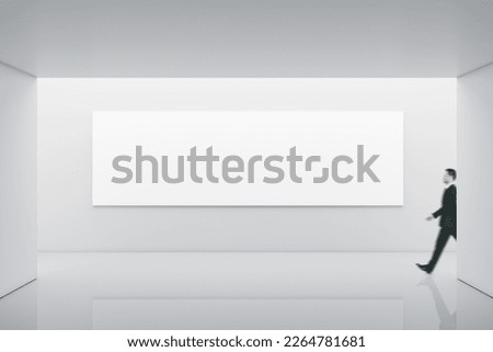 Businessman walking by long white blank poster with place for your logo or text on light illuminated wall background in abstract empty exhibition hall, mock up
