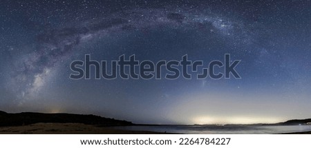 The Milky Way Panorama in the night sky with the lights from Palm Beach in Sydney on the horizon as viewed from Putty Beach on the Central Coast, NSW, Australia. Royalty-Free Stock Photo #2264784227