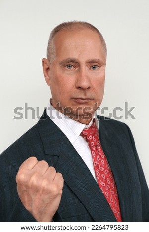 closeup portrait of elderly man in dark suit and red tie in style of Russian President Putin shows his fist against light background, concept Vladimir Putin's doubles, politics, economic sanctions Royalty-Free Stock Photo #2264795823
