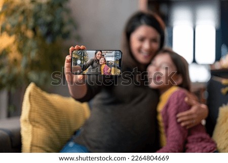 Beautiful cheerful woman and young daughter out of focus smiling looking at phone camera, recording video or shooting vlog for social media on modern smartphone at home. Focus on the phone screen.