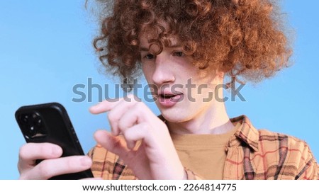 Cute young happy student on blue background looking at phone. Curly red-haired guy is crazy happy. Internet search. Technologies.
