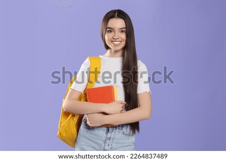 Happy teenage girl with backpack and textbooks on violet background Royalty-Free Stock Photo #2264837489