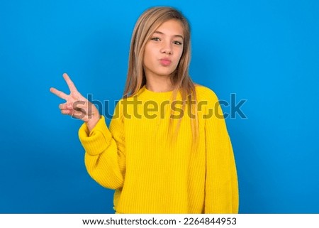 beautiful caucasian teen girl wearing yellow sweater over blue wall makes peace gesture keeps lips folded shows v sign. Body language concept
