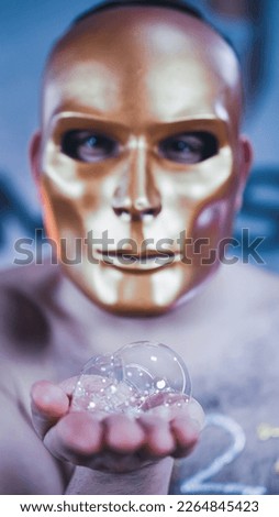 A man in a golden mask has soap bubbles on his hand.