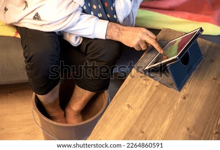 person warming cold feet in hot water at home and playing tablet, health care 