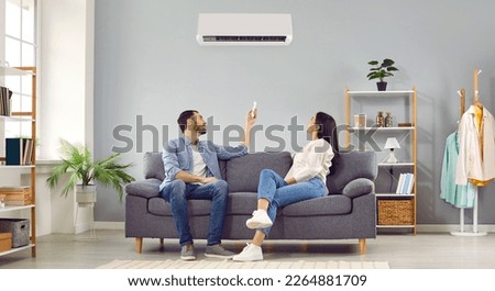 Young happy man and woman turning on air conditioner sitting on sofa at home. Smiling couple of homeowners enjoying cool conditioned air using remote resting on couch together in living room. Banner. Royalty-Free Stock Photo #2264881709