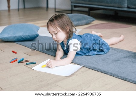 A two-year-old girl lies on the floor and draws with pencils. Living room with modern interior. 