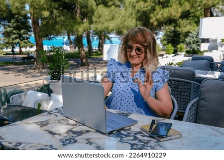 Successful, attractive, adult woman in glasses holds an online meeting in a cafe, outdoors. She salutingly mashers her hand to her followers.