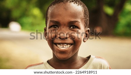 Close Up Portrait of a Shy Authentic African Boy Smiling at the Camera with a Blurred Rural Village in the Background. Black Male Kid Representing Future, Hope, and Acceptance. Documentary Footage Royalty-Free Stock Photo #2264918911
