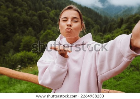 Young beautiful woman in casual wear taking selfie and blowing air kiss while standing in mountains
