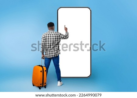 Back view of young black guy with orange suitcase traveller pushing button on phone white screen over blue studio background, interacting with touchscreen, mockup for design, copy space, collage Royalty-Free Stock Photo #2264990079