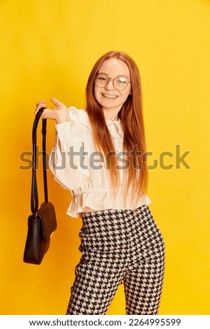 Young charming girl with red hair wearing office style clothes posing over yellow background. Concept of youth, student college life, business and education. Model looks happy, delighted