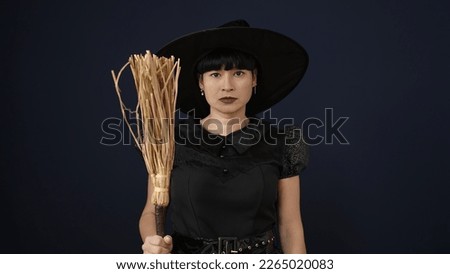 Young chinese woman wearing witch costume holding broom over isolated black background