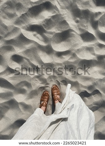 Women feet on desert beach sand. Aesthetic summer vacation background. Woman in white dress and leather sandals on the dune sand. Top view Royalty-Free Stock Photo #2265023421