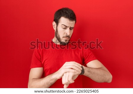 Young hispanic man wearing casual red t shirt checking the time on wrist watch, relaxed and confident  Royalty-Free Stock Photo #2265025405