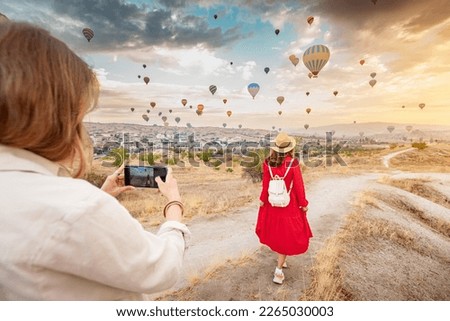 Two friends capture the beauty of Cappadocia, Turkey with their smartphones, surrounded by unique rock formations and hot air balloons. Sharing their adventure on social networks.