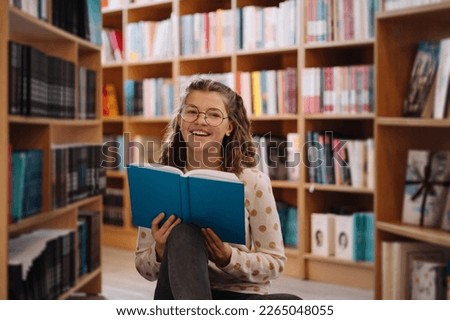 Teen girl among a pile of books. A young girl wearing glasses reads a book with shelves in the background. She is surrounded by stacks of books. Book day.