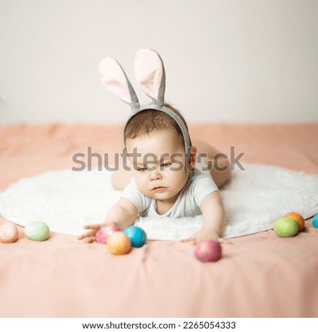 A focused infant baby is lying on the bed and reaching for a color Easter eggs, wearing bunny ears. Hunt on the Easter morning. Copy space. High quality photo