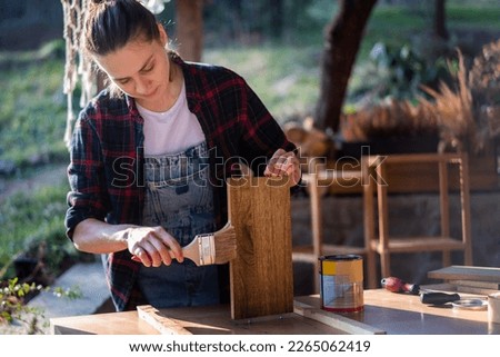 Young woman in a plaid shirt varnishing a wooden detail on a shelf on the terrace  Royalty-Free Stock Photo #2265062419