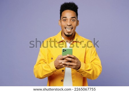 Young excited man of African American ethnicity wear yellow shirt t-shirt hold in hand use mobile cell phone isolated on plain pastel light purple background studio portrait. People lifestyle concept