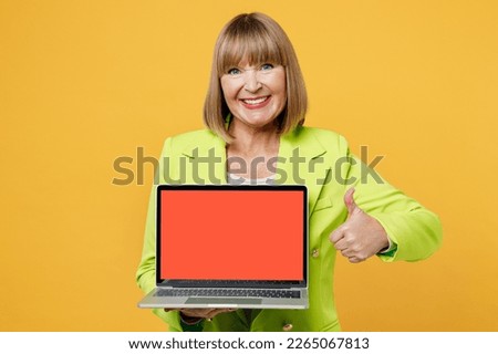 Elderly IT woman 50s years old wear green jacket white t-shirt hold use work on laptop pc computer with blank screen workspace area show thumb up isolated on plain yellow background studio portrait