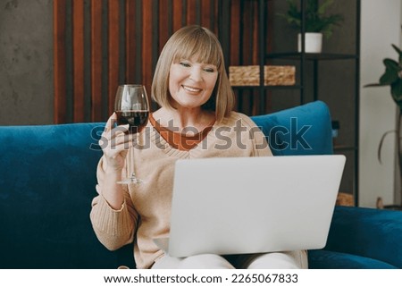 Elderly woman 50s years old wears casual clothes sits on blue sofa use laptop pc computer watch movie film drink wine stay at home flat rest relax spend free spare time in living room indoor grey wall