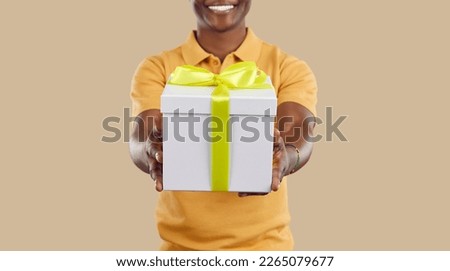Close up of festive gift box in hands of joyful dark-skinned man on beige background. White box with yellow ribbon in hands of unrecognizable smiling man. Congratulations concept. Web banner.