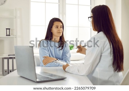 Doctor examining and consulting young beautiful female patient in hospital clinic. Woman listens to female doctor who sits with medical card near laptop on her table. Medical examination concept.