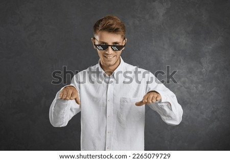 Confident young man in thug life glasses smiling, dancing, enjoying music and having fun. Happy guy in in office short and modern pixel sunglasses doing funny dance moves isolated on grey background