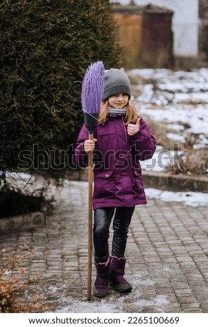 A little diligent girl, a child with a broom in her hands, is satisfied with her work done outdoors in nature. Photography, portrait, childhood concept.