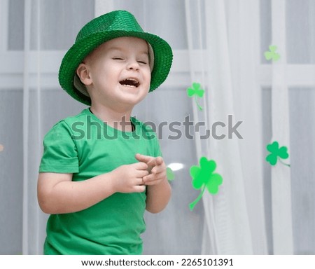St. Patrick's Day concept. A little cheerful boy in a shiny green top hat and green clothes laughs against the background of a window decorated with a three-leaf clover made of paper