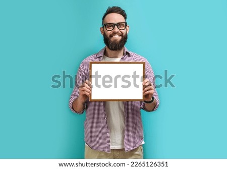 A handsome guy with a beard in eyeglasses proudly holds in his hands a frame with an empty space for your diploma, award or text. Recognition for good work Royalty-Free Stock Photo #2265126351