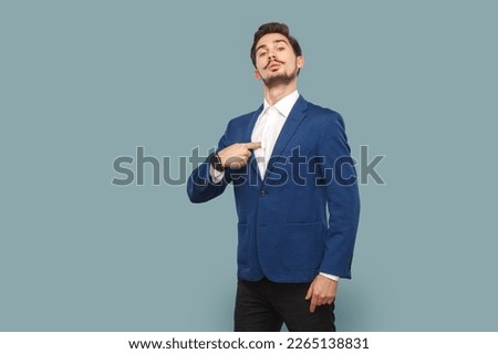 Portrait of self confident proud man with mustache standing pointing at himself, looking at camera with pride, wearing white shirt and jacket. Indoor studio shot isolated on light blue background. Royalty-Free Stock Photo #2265138831