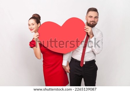 Portrait of smiling satisfied man in white shirt and woman in red dress standing together, peeping out from big heart, expressing love. Indoor studio shot isolated on gray background.