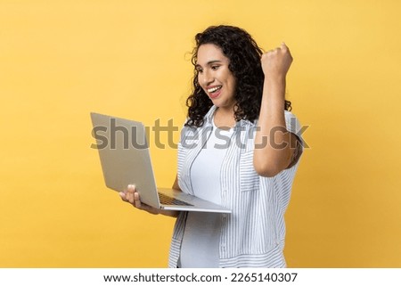 Portrait of extremely happy woman with dark wavy hair working on laptop and looking at display, clenched fist, celebrating her winning. Indoor studio shot isolated on yellow background.