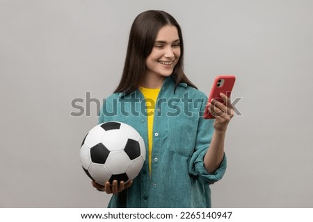 Smiling delighted woman standing holding soccer ball and using smart phone, expressing positive emotions, wearing casual style jacket. Indoor studio shot isolated on gray background.