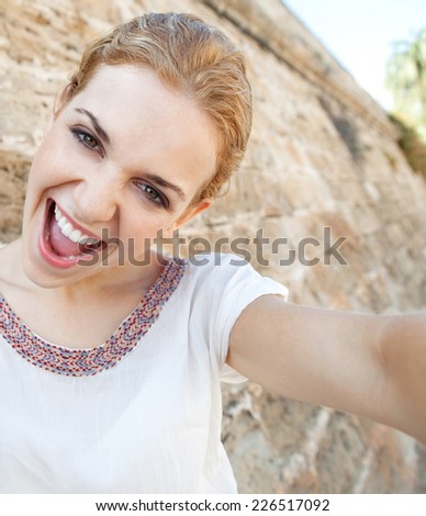 Portrait of a joyful young tourist woman visiting a monument with a textured old stone wall, using a smartphone device to take a selfie self portrait picture of herself, networking. Travel technology.