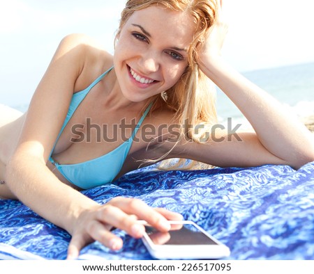 Beautiful young tourist teenager woman on holiday relaxing on a beach towel in a sandy sunny summer beach looking at photos on a smartphone device with touch screen. Travel technology outdoors.
