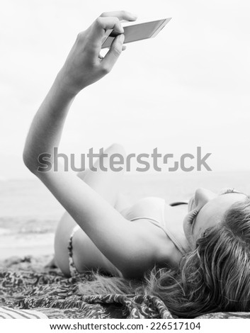 Black and white over head rear view of a young tourist woman on holiday relaxing laying on a beach holding a smartphone device up in her hands with touch screen. Travel vacation technology.