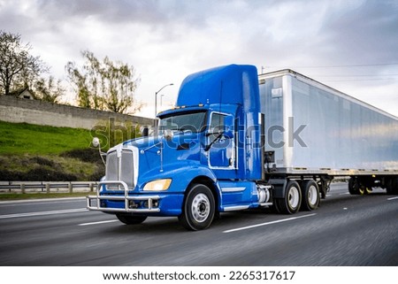 Day cab blue big rig industrial semi trucks tractor with roof spoiler and turn on headlight transporting commercial cargo in dry van semi trailer running on the wide highway road at twilight time Royalty-Free Stock Photo #2265317617
