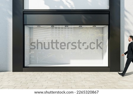 Businessman walking through sunlit empty showcase with light wall background behind window with space for product presentation in modern business center building outdoors, mock up Royalty-Free Stock Photo #2265320867