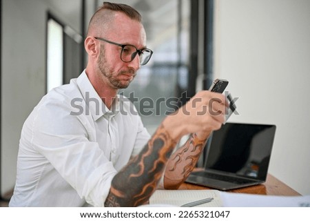 Side view, Confused and stressed Caucasian man with glasses and tattoo on his arms sits at his desk having a problem on his mobile banking app. credit card or debit card payment concept