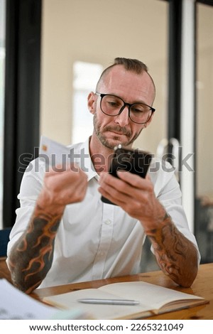 Portrait, a Caucasian businessman sits at his desk using mobile banking application to pay his online bills, holding a credit card and smartphone.