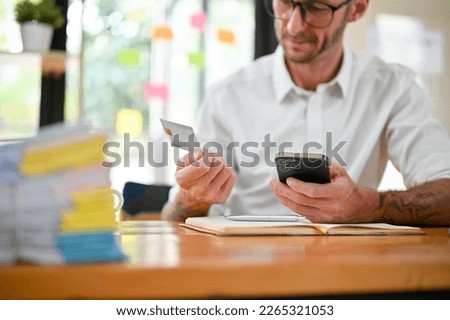 Cropped image of a Caucasian businessman or male accountant sits at his desk using mobile banking application to pay his online bills, holding a credit card and smartphone.