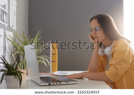 Horizontal shot of calm beautiful woman dressed in yellow shirt using laptop computer and sitting at table in office, looking at notebook monitor, learning information.