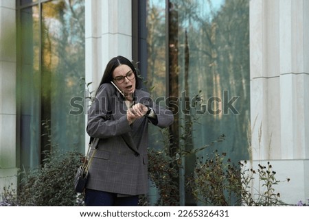 Emotional woman checking time while talking on smartphone outdoors. Being late concept Royalty-Free Stock Photo #2265326431