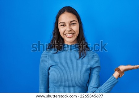 Young brazilian woman standing over blue isolated background smiling cheerful presenting and pointing with palm of hand looking at the camera. 