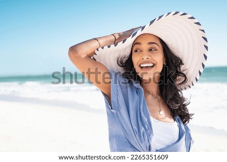 Carefree woman with white and blue straw hat walking at beach. Young smiling latin woman on vacation enjoying sea breeze. Attractive hispanic girl relaxing at beach with the sea behind.